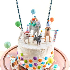 May include: A white cake decorated with colorful sprinkles and a border of colorful chocolate candies.  The cake is topped with a variety of dog figurines wearing party hats and holding balloons and gifts.  The cake is on a wooden cake stand with a banner that reads "Happy Birthday".