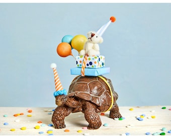 May include: A brown tortoise wearing a blue and orange party hat carries a white teddy bear wearing a blue and white party hat. The teddy bear is sitting on top of a blue and white gift box with a blue ribbon. The tortoise is surrounded by colorful confetti on a wooden surface.