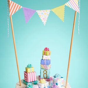May include: A white cake decorated with a stack of colorful gift boxes and a paper banner with pastel colored triangles. The cake is topped with a number 8.