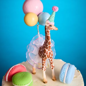 May include: A decorative cake topper with a giraffe wearing a party hat and holding balloons. The giraffe is brown with white spots, and the cake is decorated with macarons and sprinkles. The balloons are pink, yellow, and blue.