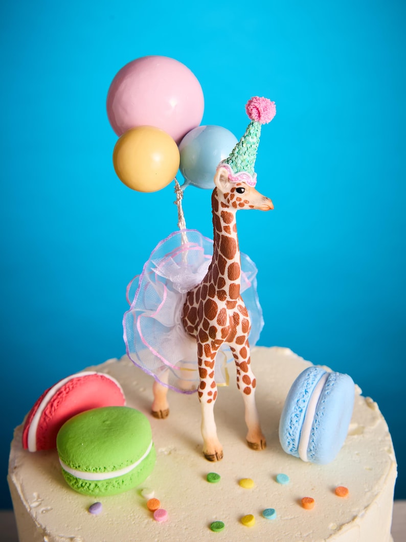 May include: A decorative cake topper with a giraffe wearing a party hat and holding balloons. The giraffe is brown with white spots, and the cake is decorated with macarons and sprinkles. The balloons are pink, yellow, and blue.