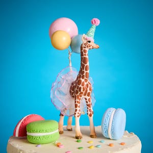 May include: A decorative cake topper with a giraffe wearing a party hat and holding balloons. The giraffe has brown and white markings and a tutu. The cake base has colourful macarons in red, green, and blue, plus sprinkles.