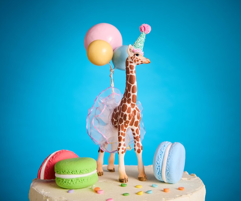 May include: A decorative cake topper with a giraffe wearing a party hat and holding balloons. The giraffe has brown and white markings and a tutu. The cake base has colourful macarons in red, green, and blue, plus sprinkles.