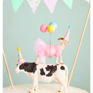 May include: A whimsical cake topper featuring a cow with black and white markings, topped by a pink goat wearing a tutu and party hat. The goat holds balloons. A banner with pastel flags hangs in the background.