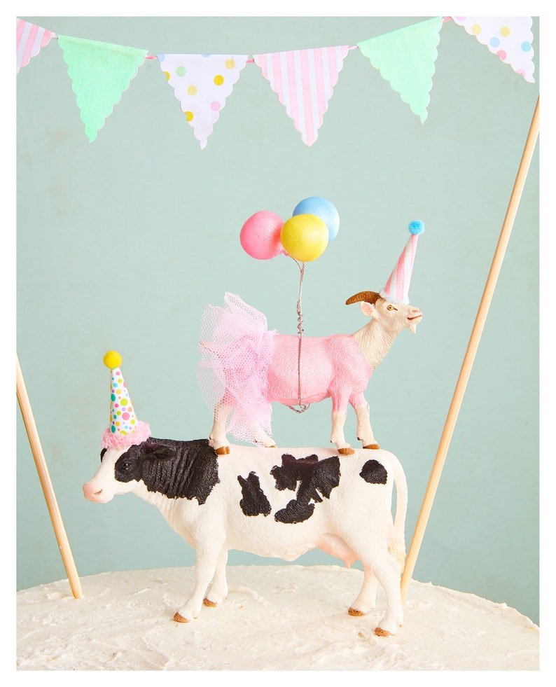 May include: A whimsical cake topper featuring a cow with black and white markings, topped by a pink goat wearing a tutu and party hat. The goat holds balloons. A banner with pastel flags hangs in the background.