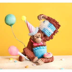 May include: A brown monkey figurine wearing a blue and white striped party hat, a blue and white bow tie, and a blue and white striped vest. The monkey is holding a blue and white gift box and a pink donut. There are pink and green balloons and confetti on the table.