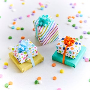 May include: Four small gift boxes wrapped in colorful paper with ribbons and bows. The boxes are on a white surface with colorful sprinkles scattered around them.