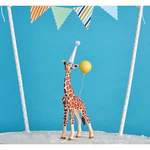 May include: A whimsical cake topper featuring a giraffe wearing a party hat and holding a yellow balloon. The giraffe has brown spots and is set against a blue background with colorful pennant flags. The topper is designed for a birthday cake.