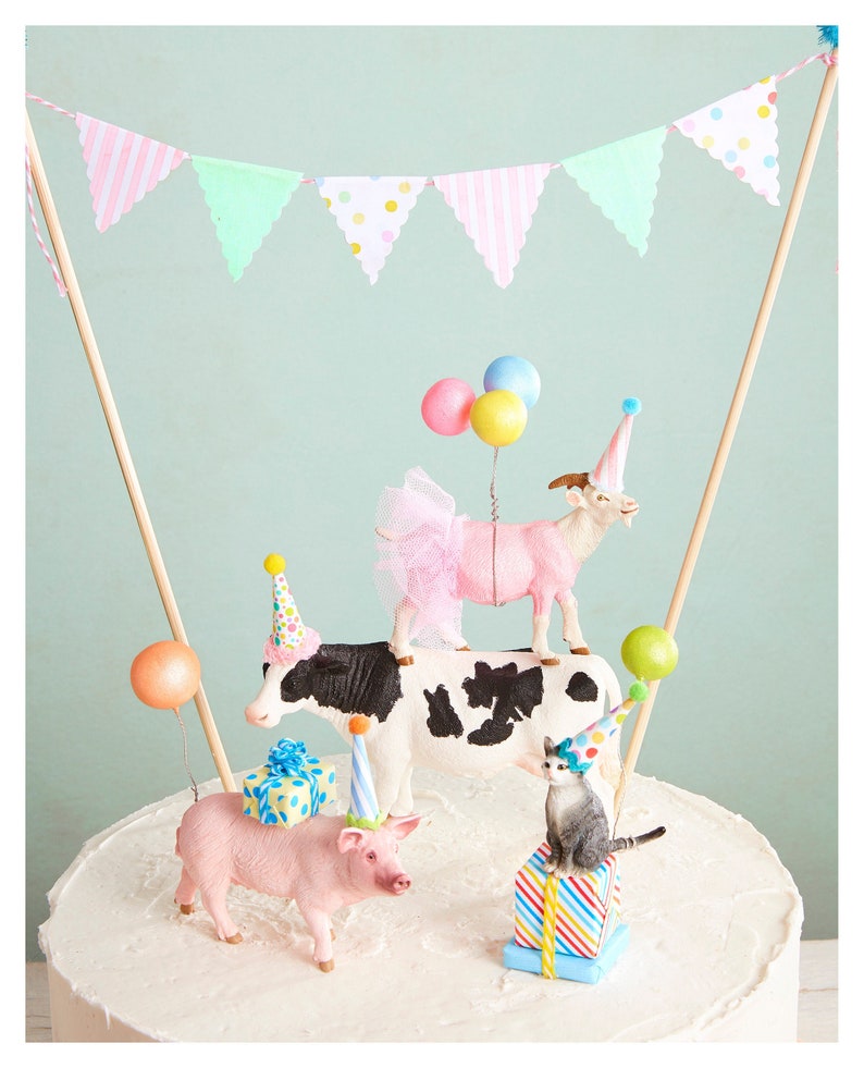 May include: A whimsical cake topper featuring a cow, goat, pig, and cat, each wearing a party hat. The goat wears a pink tutu. Colorful balloons and a banner with pink and green flags complete the festive design. The cake topper is designed for a birthday celebration.