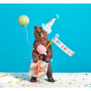 May include: A brown bear figurine wearing a colorful party hat and holding a gift box and a banner that says "Happy Birthday".