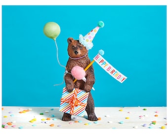 May include: A brown bear figurine wearing a colorful party hat and holding a gift box and a banner that says "Happy Birthday".