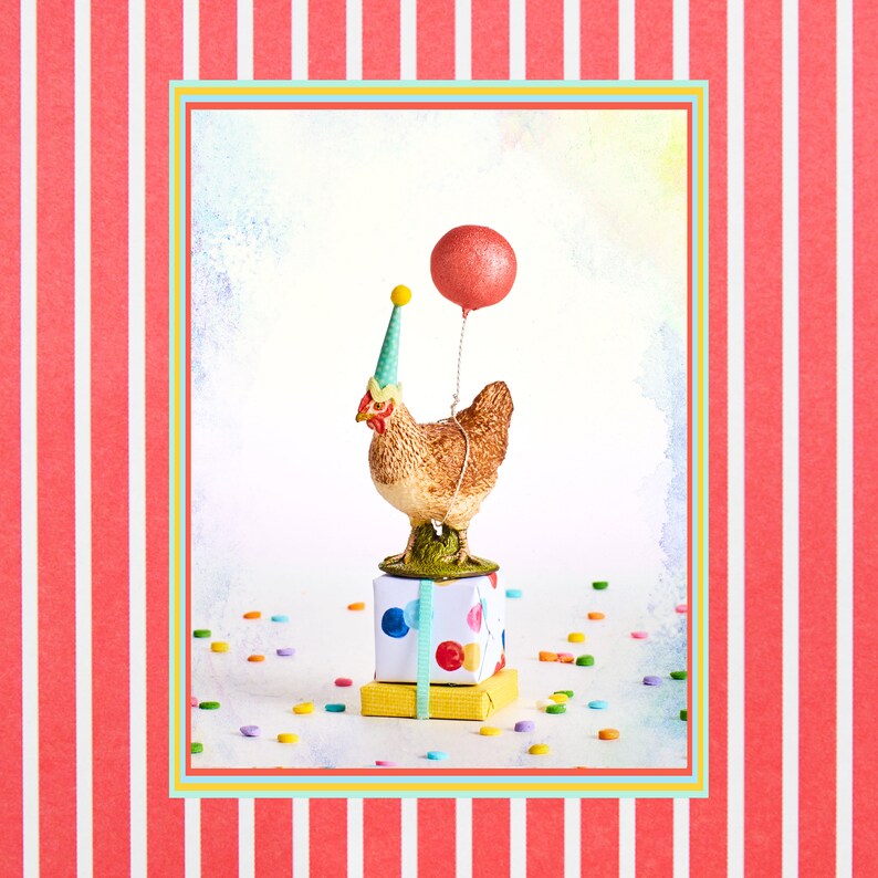 May include: A whimsical figurine of a brown chicken wearing a party hat and holding a red glitter balloon. The chicken stands atop a stack of wrapped gifts with colorful polka dots and ribbons. Confetti is scattered around the base.