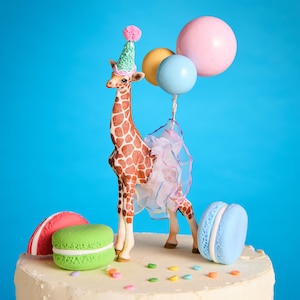 May include: A novelty cake topper of a giraffe wearing a party hat and holding balloons. The giraffe has brown and tan markings and a tulle skirt. The cake is decorated with macarons and sprinkles.