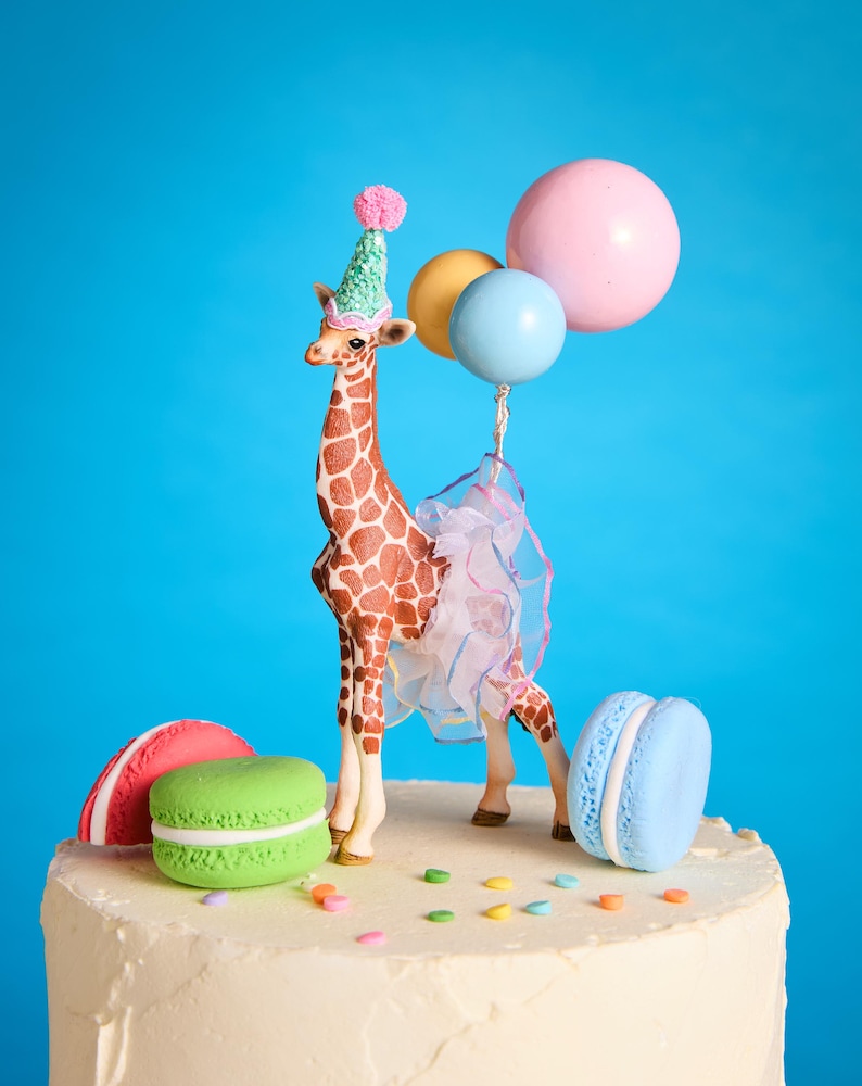 May include: A novelty cake topper of a giraffe wearing a party hat and holding balloons. The giraffe has brown and tan markings and a tulle skirt. The cake is decorated with macarons and sprinkles.