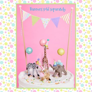 May include: A white cake decorated with colorful sprinkles and topped with a giraffe, zebra, lion, and elephant figurines wearing party hats. A pink and white bunting banner with the text "Banners sold separately" hangs above the cake.