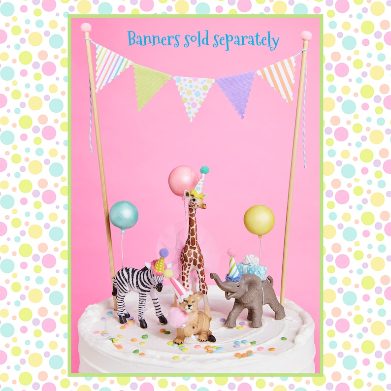 May include: A white cake decorated with colorful sprinkles and topped with a giraffe, zebra, lion, and elephant figurines wearing party hats. A pink and white bunting banner with the text "Banners sold separately" hangs above the cake.