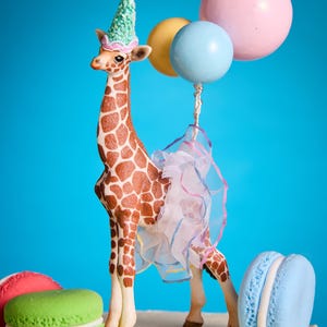 May include: A decorative giraffe figurine with a party hat and tutu, holding balloons. The giraffe has brown markings and is set against a blue backdrop, with colorful macarons nearby.