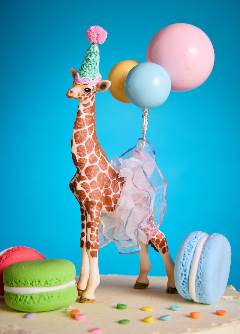May include: A decorative giraffe figurine with a party hat and tutu, holding balloons. The giraffe has brown markings and is set against a blue backdrop, with colorful macarons nearby.