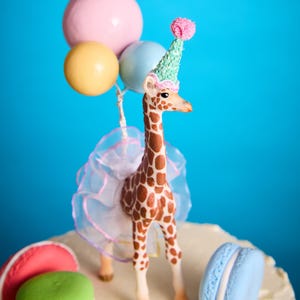 May include: A decorative cake topper of a giraffe wearing a party hat and tutu, holding balloons. The giraffe has brown spots and is surrounded by macarons. The balloons are pink, yellow, and blue. The cake is decorated with macarons.