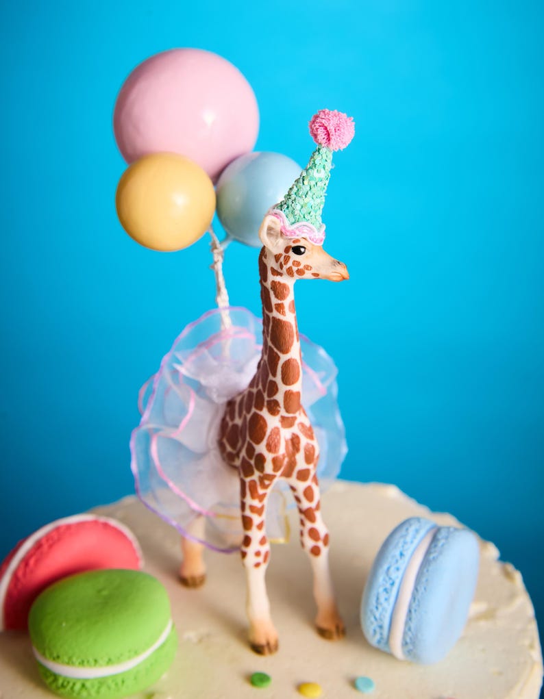 May include: A decorative cake topper of a giraffe wearing a party hat and tutu, holding balloons. The giraffe has brown spots and is surrounded by macarons. The balloons are pink, yellow, and blue. The cake is decorated with macarons.