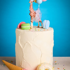 May include: A white frosted cake decorated with a giraffe figurine wearing a party hat and holding balloons. The cake is adorned with colourful macarons, an ice cream cone, and a doughnut. Sprinkles are scattered around the base.