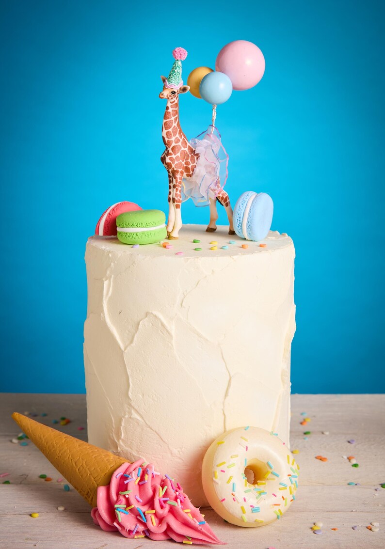 May include: A white frosted cake decorated with a giraffe figurine wearing a party hat and holding balloons. The cake is adorned with colourful macarons, an ice cream cone, and a doughnut. Sprinkles are scattered around the base.