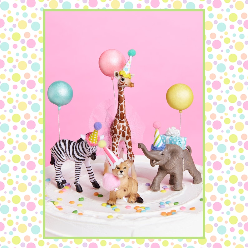 May include: A white cake decorated with colorful sprinkles and animal figurines. A giraffe, zebra, elephant, and lion are wearing party hats. Balloons are floating above the cake.