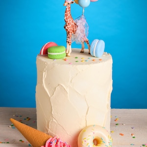 May include: A celebratory cake featuring a giraffe figure with a party hat and balloons. The cake is decorated with macarons, a pink ice cream cone, and a donut, with colourful sprinkles. The background is a vibrant blue.