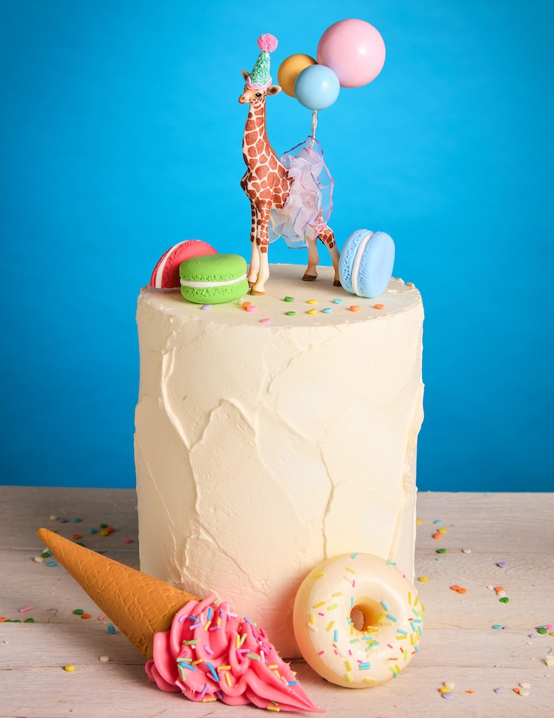 May include: A celebratory cake featuring a giraffe figure with a party hat and balloons. The cake is decorated with macarons, a pink ice cream cone, and a donut, with colourful sprinkles. The background is a vibrant blue.