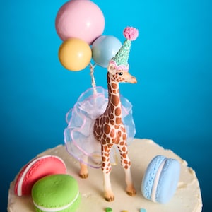 May include: A decorative cake topper with a giraffe wearing a party hat and tutu, holding balloons. The cake is adorned with macarons and sprinkles. The background is a vibrant blue.