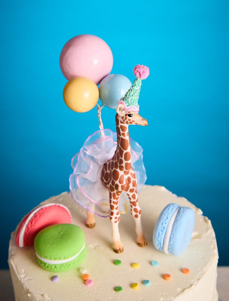 May include: A decorative cake topper with a giraffe wearing a party hat and tutu, holding balloons. The cake is adorned with macarons and sprinkles. The background is a vibrant blue.