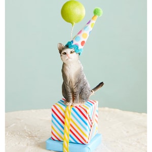 May include: A figurine of a gray and white cat wearing a party hat and sitting atop wrapped presents. The cat holds a green balloon. The presents are wrapped in colorful striped paper and blue paper, tied with yellow ribbon.