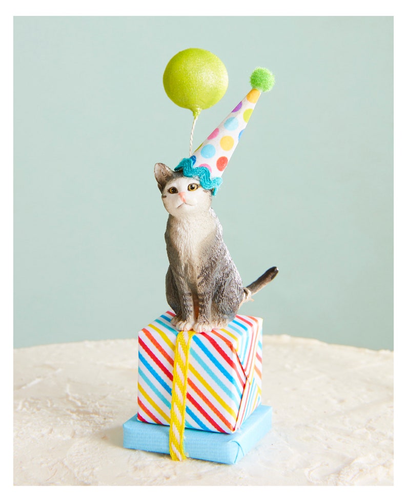 May include: A figurine of a gray and white cat wearing a party hat and sitting atop wrapped presents. The cat holds a green balloon. The presents are wrapped in colorful striped paper and blue paper, tied with yellow ribbon.