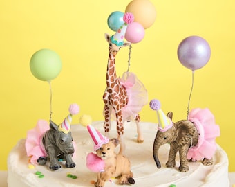May include: A white cake decorated with colorful sprinkles and animal figurines. The figurines include a giraffe, a rhino, a lion, and an elephant. Each animal is wearing a pink tutu and a party hat. There are also colorful balloons on the cake.
