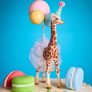 May include: A decorative cake topper of a giraffe wearing a party hat and tutu, holding balloons. The cake is decorated with colourful macarons and sprinkles, set against a bright blue background. A fun, celebratory theme.
