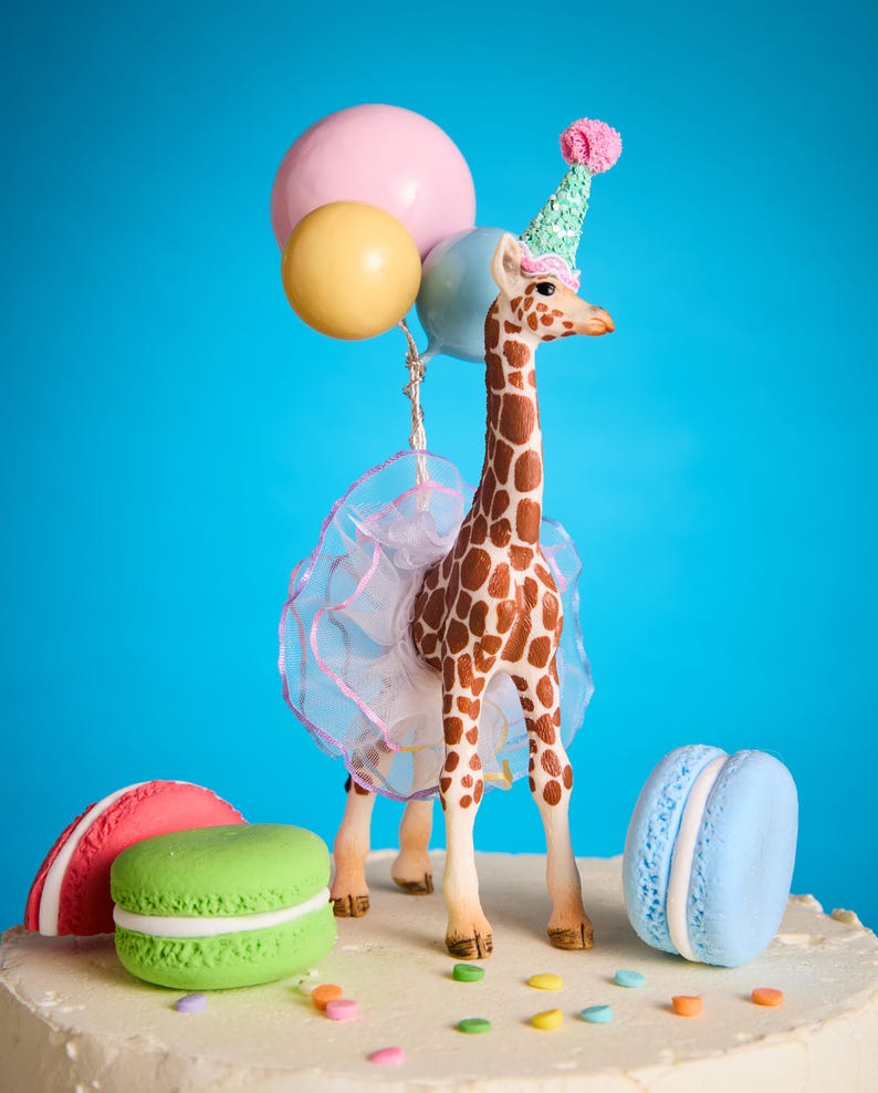 May include: A decorative cake topper of a giraffe wearing a party hat and tutu, holding balloons. The cake is decorated with colourful macarons and sprinkles, set against a bright blue background. A fun, celebratory theme.