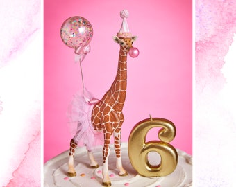 May include: A pink and brown giraffe cake topper wearing a pink tutu and a party hat, holding a pink balloon. The giraffe is standing next to a gold number six candle.