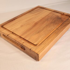 May include: A rectangular wooden cutting board with a juice groove and a handle. The board is made of light brown wood and has a natural wood grain pattern. A logo is visible on the side of the board.