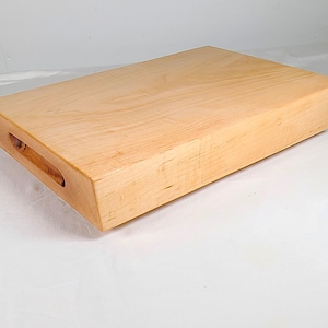 May include: A thick, rectangular light-colored wooden cutting board with a smooth, polished finish. The board features a beveled edge design and a carved handle cutout on the side, showcasing the natural grain patterns of the light wood.