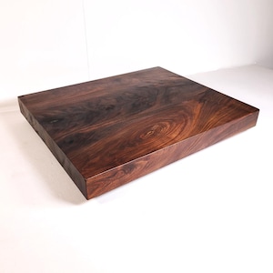May include: A square, solid wood cutting board with a rich, dark brown color and a polished finish. The wood grain is visible, creating a natural, swirling pattern. The board is thick and appears to be made from a single piece of wood.