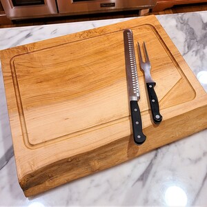 May include: A large, rectangular wooden cutting board with a natural wood grain finish. The board has a raised edge and a groove around the perimeter. Two silver kitchen knives with black handles are placed on the board.