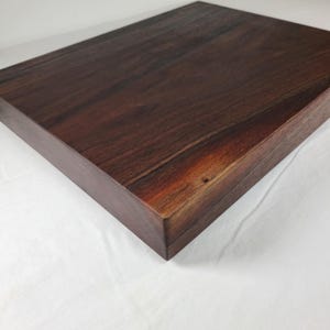 May include: A rectangular, solid wood cutting board with a rich, dark brown color and visible wood grain. The edges are clean and square, and the surface appears smooth and well-finished. The board is set against a white background.