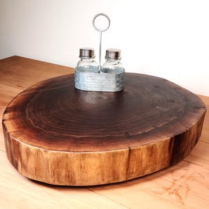 May include: A round wooden serving tray made from a tree slice with a natural wood grain pattern. The tray has a metal salt and pepper shaker holder with a ring on top.