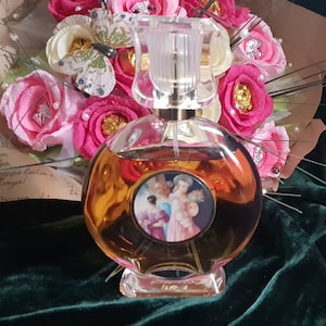 May include: A clear glass bottle of perfume with a gold cap and a label featuring a vintage illustration of women in a garden. The bottle is sitting on a green velvet surface.