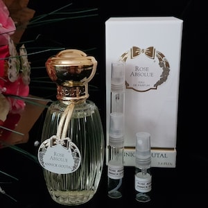 Op de afbeelding: Een doorzichtige glazen fles Rose Absolue parfum van Annick Goutal. De fles heeft een gouden dop en een gouden etiket met de naam van het parfum. De fles staat op een zwarte ondergrond naast een kleinere fles van hetzelfde parfum en een witte doos met de naam van het parfum erop.