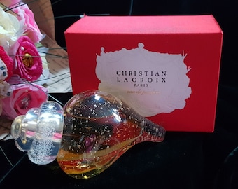 May include: A clear glass perfume bottle shaped like a seashell with gold flecks inside. The bottle is sitting on a dark green surface next to a red box with the words "Christian Lacroix Paris eau de parfum" printed on it.