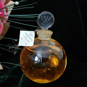 May include: A clear glass bottle of perfume with a gold liquid and a star-shaped design. The bottle has a clear glass stopper with a silver letter "W" on top. The label on the bottle reads "Eau de Toilette "Je Reviens" 80g Worth Paris France".