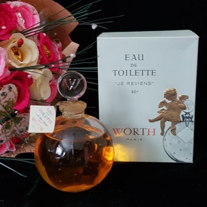 May include: A vintage bottle of Worth "Je Reviens" Eau de Toilette perfume. The bottle is clear glass with a gold-colored stopper and a W on top. The bottle is filled with a golden liquid. The box is white with gold lettering and a gold cherub on the front. The box says "Eau de Toilette "Je Reviens" 80&deg; Worth Paris"