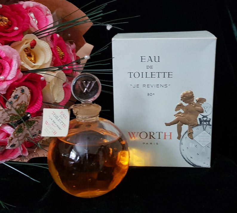 May include: A vintage bottle of Worth "Je Reviens" Eau de Toilette perfume. The bottle is clear glass with a gold-colored stopper and a W on top. The bottle is filled with a golden liquid. The box is white with gold lettering and a gold cherub on the front. The box says "Eau de Toilette "Je Reviens" 80&deg; Worth Paris"