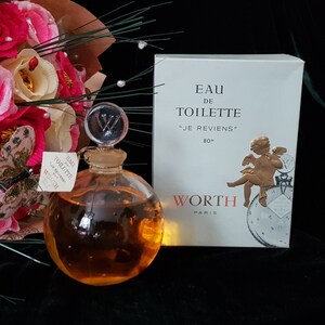 May include: A vintage glass bottle of perfume with a gold cap and a gold label that reads "Eau de Toilette "Je Reviens" 80&deg; Worth Paris". The bottle is round and clear, and the liquid inside is a light amber color. The bottle is sitting on a black surface.